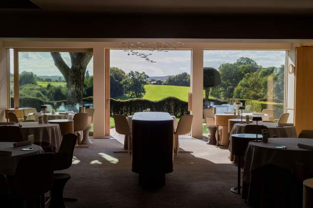 Une salle de restaurant lumineuse, dotée de larges baies vitrées donnant sur la campagne, avec des tables élégamment dressées.